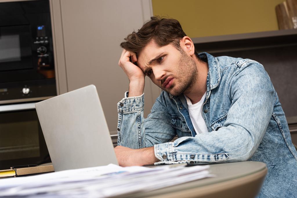 Selective focus of upset man looking at laptop near papers on kitchen table  - Photo, Image