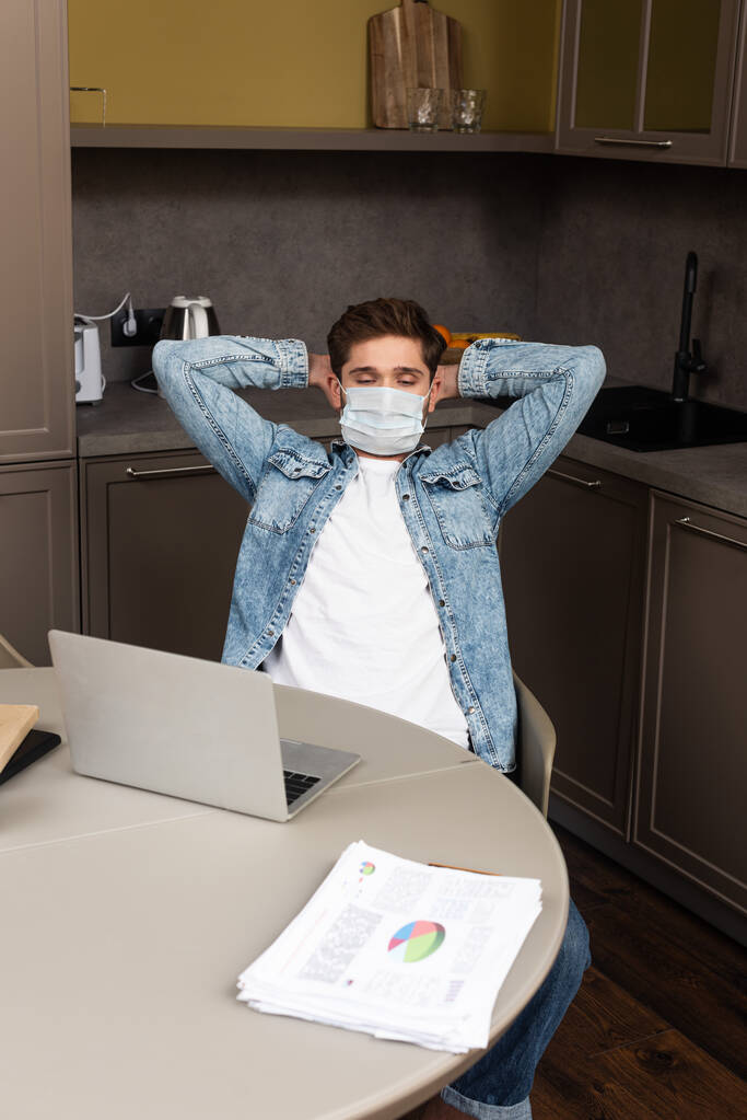 Selective focus of freelancer in medical mask sitting at kitchen table with laptop and papers  - Photo, Image