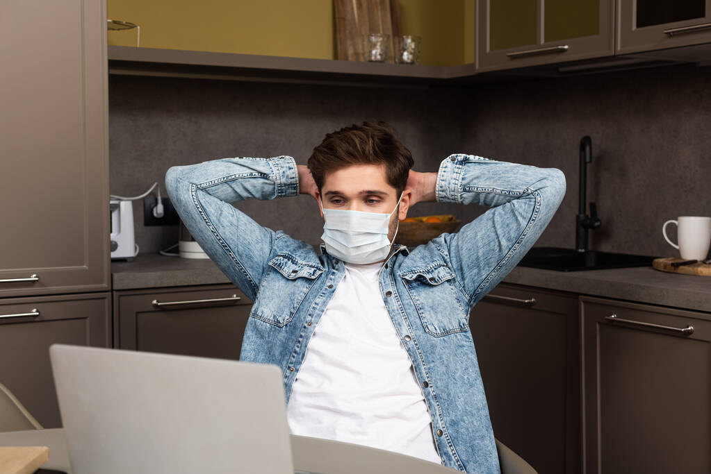 Selective focus of freelancer in medical mask looking at laptop on kitchen table  - Photo, Image