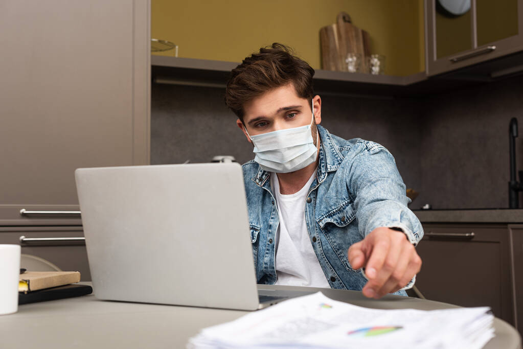 Selective focus of man in medical mask taking papers near laptop on table in kitchen  - Photo, Image