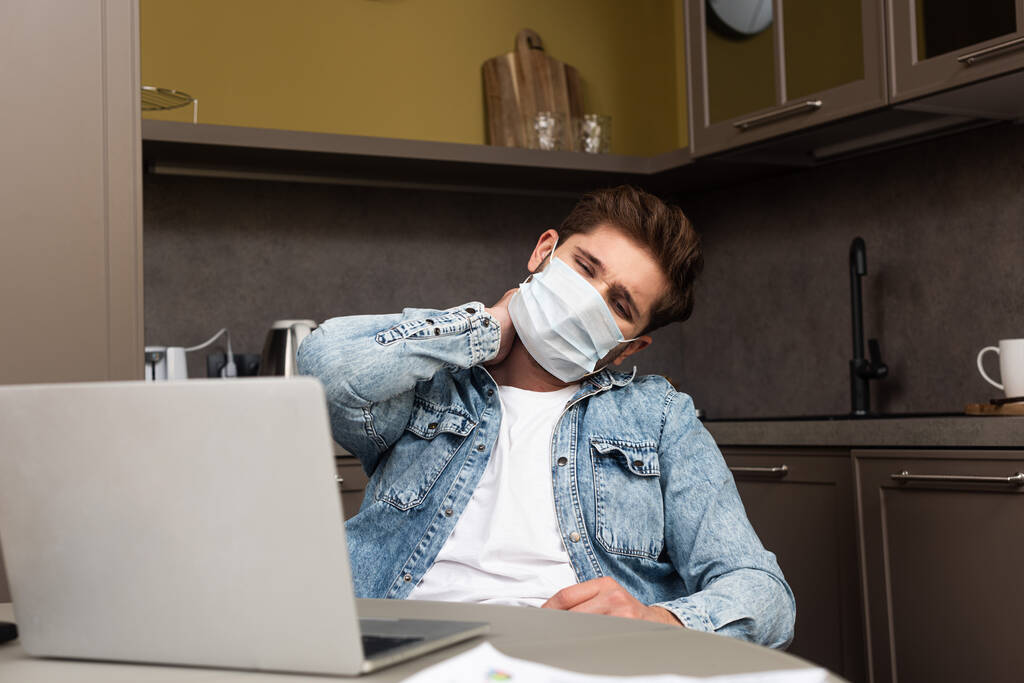 Selektywne skupienie się na wyczerpanym freelancerze w masce medycznej siedzącej obok laptopa na stole kuchennym  - Zdjęcie, obraz