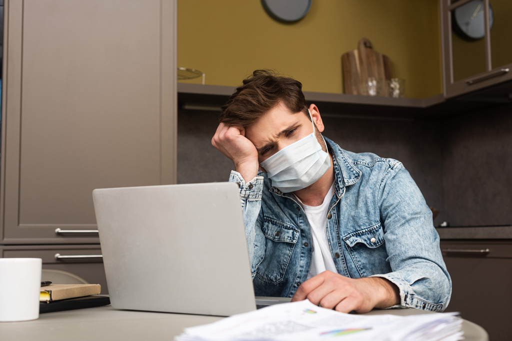紙やノートパソコンとテーブルに座って医療マスクで悲しいフリーランスの選択的な焦点  - 写真・画像