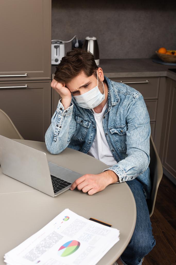 Zdenerwowany telepracownik w masce medycznej patrząc na laptopa w pobliżu dokumentów z wykresami na stole kuchennym  - Zdjęcie, obraz