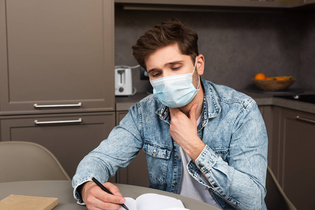 Orvosi maszkos férfi megérinti a nyakát, miközben jegyzetfüzetre ír a konyhaasztalon.  - Fotó, kép