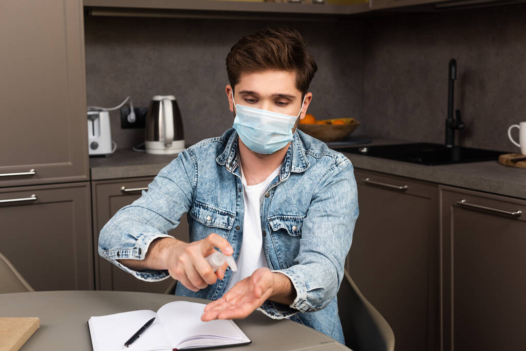 Человек в медицинской маске с помощью дезинфицирующего средства для рук возле ноутбука на кухонном столе
  - Фото, изображение