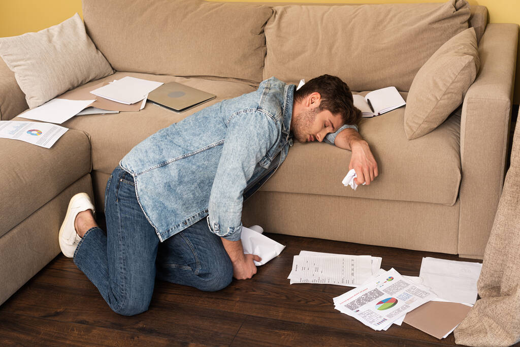 Вид сбоку на красивого мужчину спящего, держащего сгущенную бумагу рядом с ноутбуком на диване
  - Фото, изображение