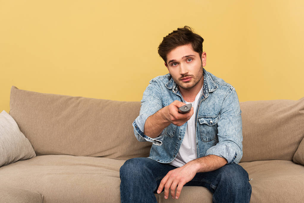 Hombre guapo sosteniendo mando a distancia mientras está sentado en el sofá
  - Foto, Imagen