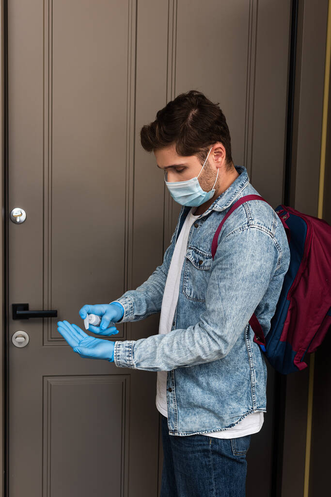 Vista lateral do homem em luvas de látex e máscara médica usando higienizador de mão perto da porta em casa
  - Foto, Imagem