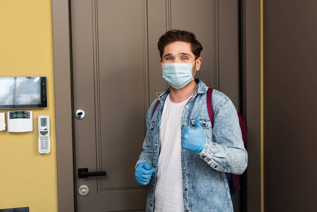 Homme en masque médical et gants en latex montrant comme geste près de la porte dans le couloir
  - Photo, image