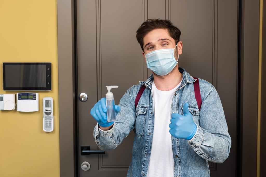 Mann mit medizinischer Maske und Latexhandschuhen zeigt Daumen hoch und hält Händedesinfektionsmittel in der Nähe des Körpers zu Hause  - Foto, Bild