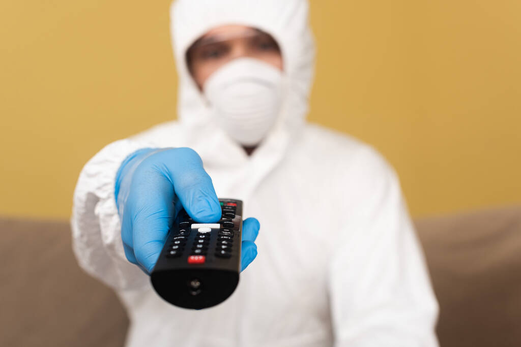 Επιλεκτική εστίαση του ανθρώπου στο γάντι λατέξ, τη στολή και την ιατρική μάσκα κρατώντας τηλεχειριστήριο στο σπίτι  - Φωτογραφία, εικόνα