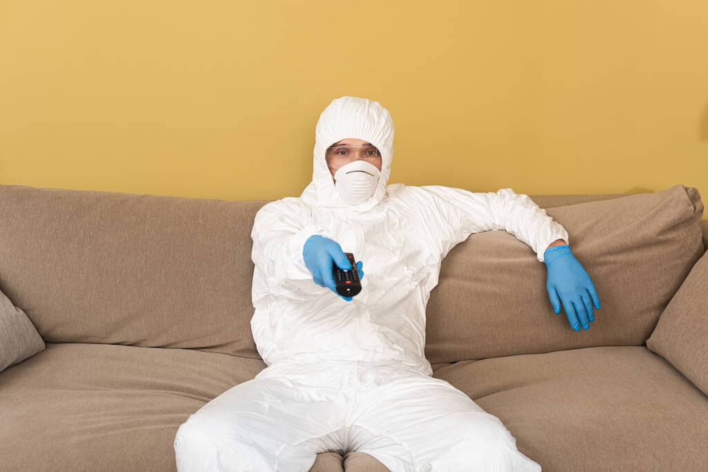 Hombre con máscara médica, gafas de seguridad y traje de materiales peligrosos que sostiene el mando a distancia en el sofá
  - Foto, Imagen