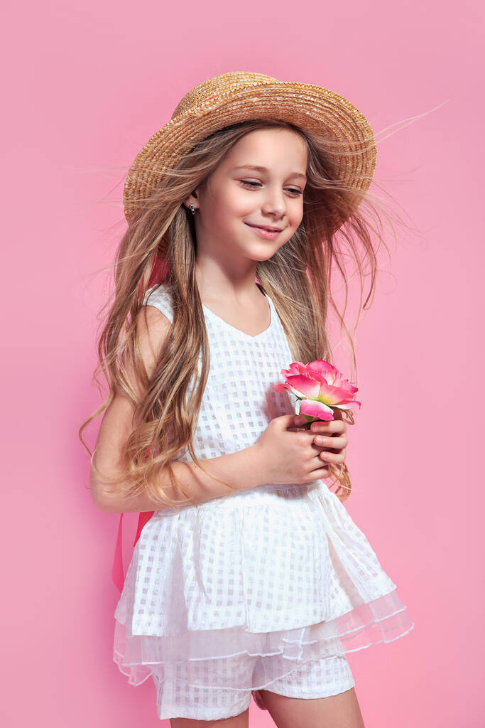 Μικρό όμορφο κορίτσι με λουλούδι ντυμένο με λευκό φόρεμα και ψάθινο καπέλο σε ροζ φόντο - Φωτογραφία, εικόνα