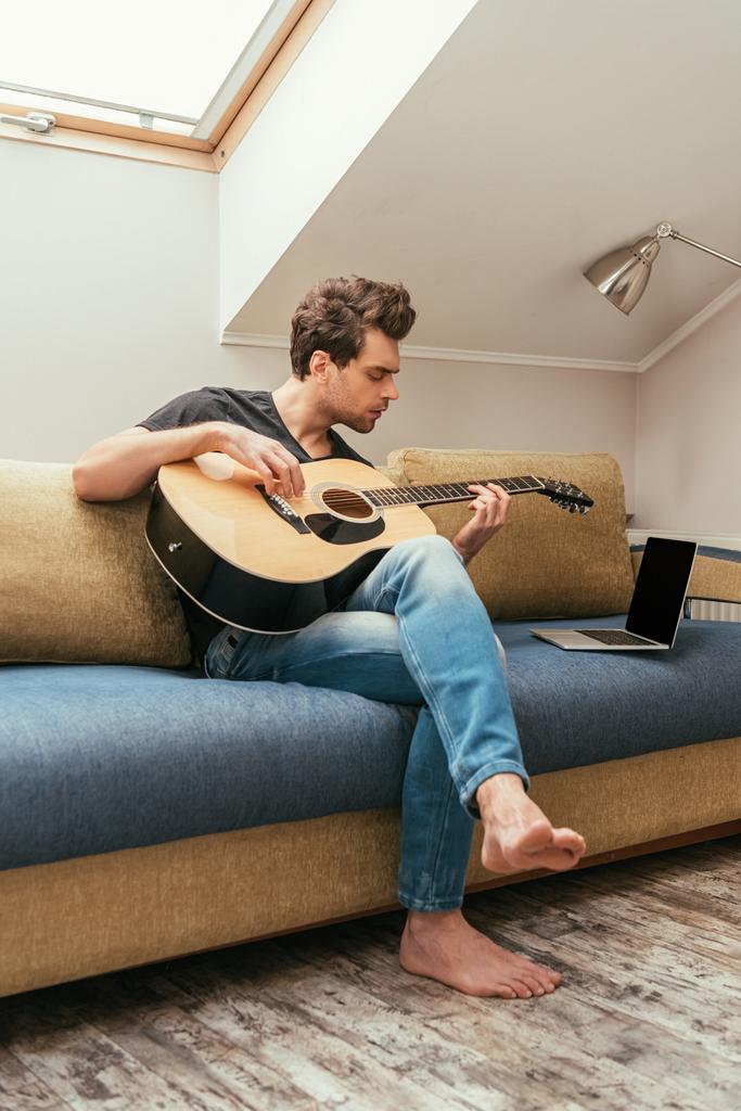 atento joven tocando la guitarra mientras está sentado en el sofá cerca de la computadora portátil con pantalla en blanco
 - Foto, Imagen