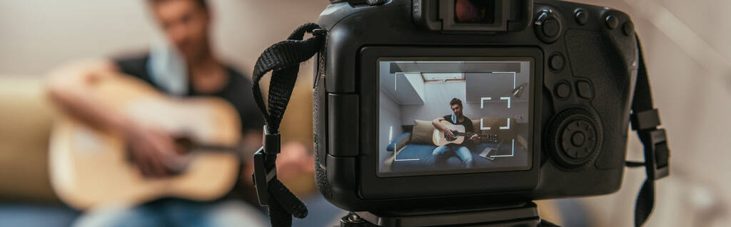foco seletivo do jovem vlogger tocando guitarra enquanto olha para a câmera digital, imagem horizontal
 - Foto, Imagem
