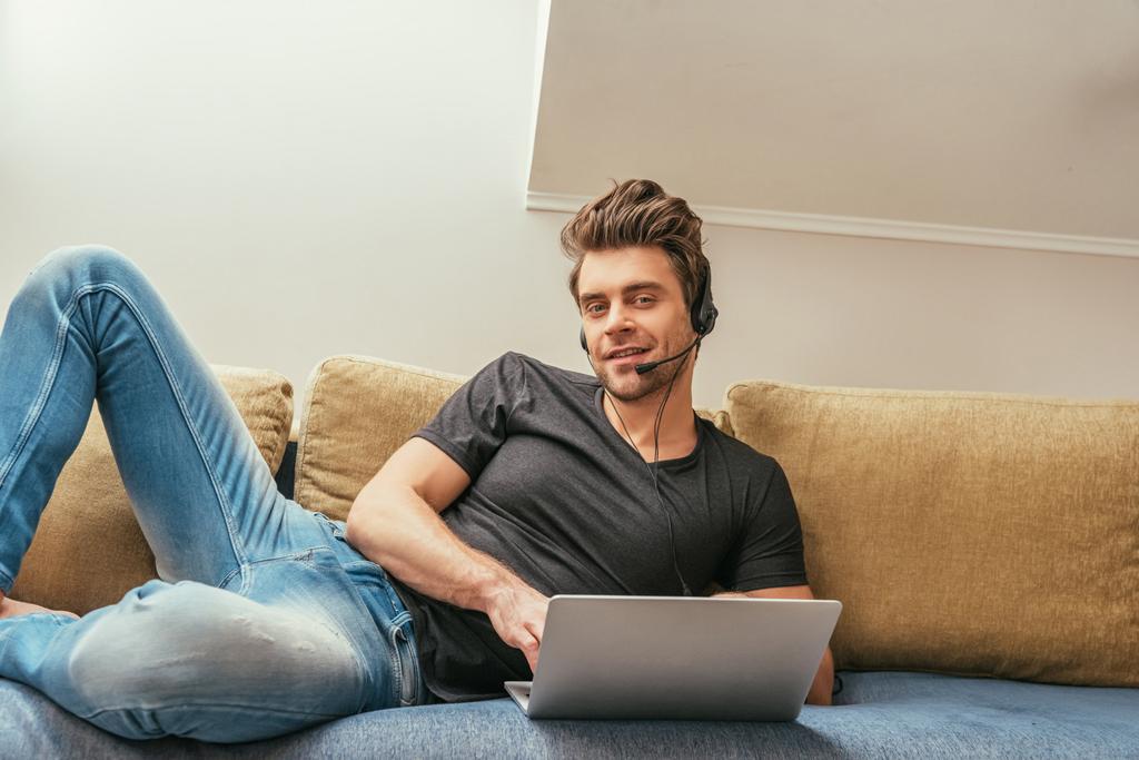 hombre guapo en auriculares tumbado en el sofá cerca de la computadora portátil y sonriendo a la cámara
 - Foto, imagen