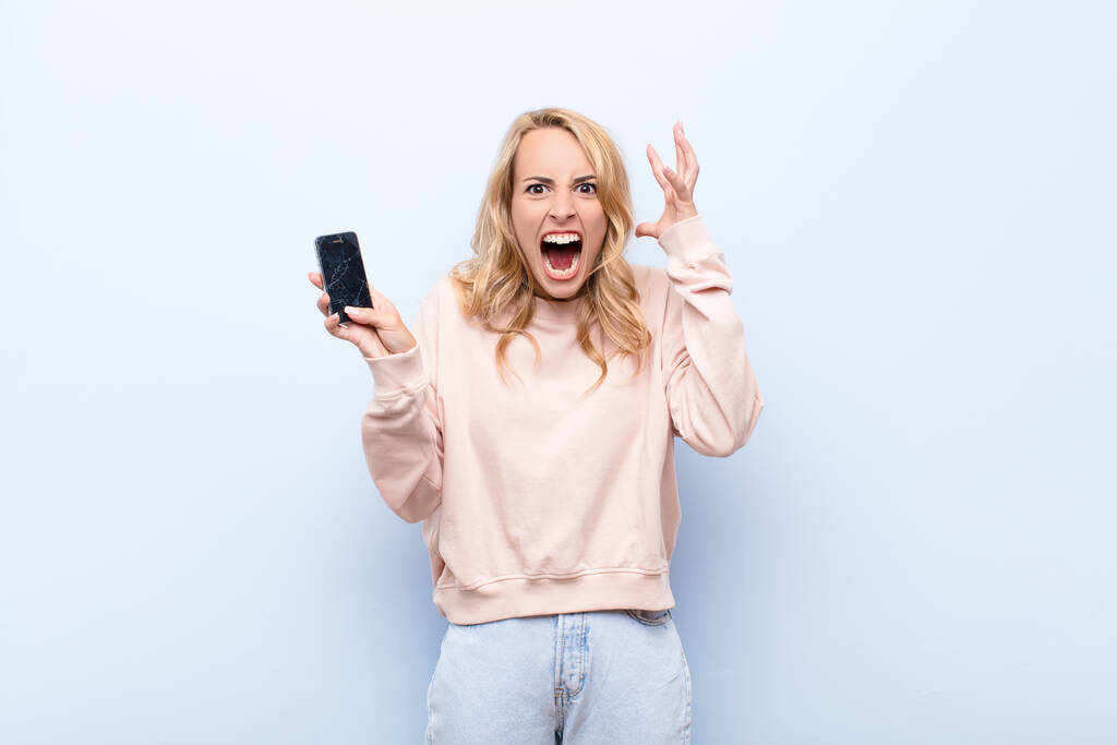 νεαρή ξανθιά γυναίκα ουρλιάζει με τα χέρια ψηλά στον αέρα, αισθάνεται έξαλλος, απογοητευμένος, αγχωμένος και αναστατωμένος χρησιμοποιώντας ένα smartphone - Φωτογραφία, εικόνα