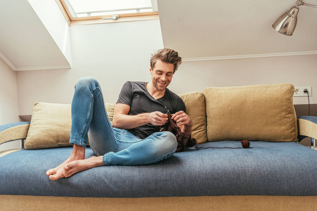χαρούμενος νεαρός άνδρας πλέξιμο, ενώ κάθεται στον καναπέ στο σπίτι στη σοφίτα δωμάτιο - Φωτογραφία, εικόνα