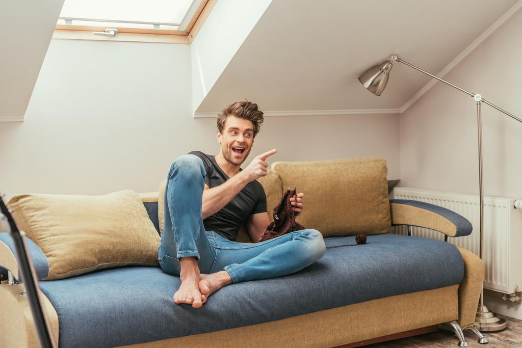 aufgeregter junger Mann zeigt mit Finger beim Stricken auf Sofa zu Hause - Foto, Bild