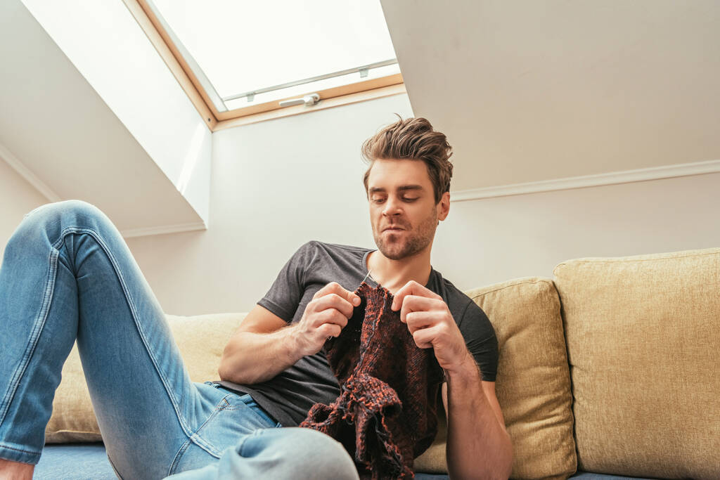низкий угол зрения на недовольного человека вязания во время сидения на диване в мансарде
 - Фото, изображение