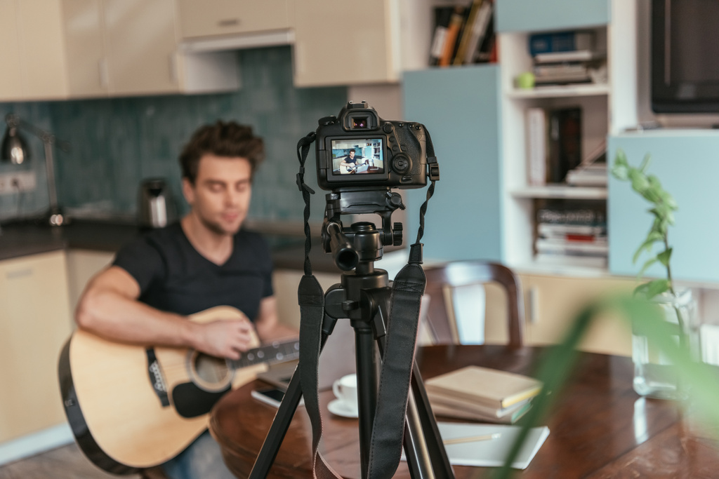 selective focus of young vlogger playing guitar in kitchen near digital camera on tripod - Photo, Image