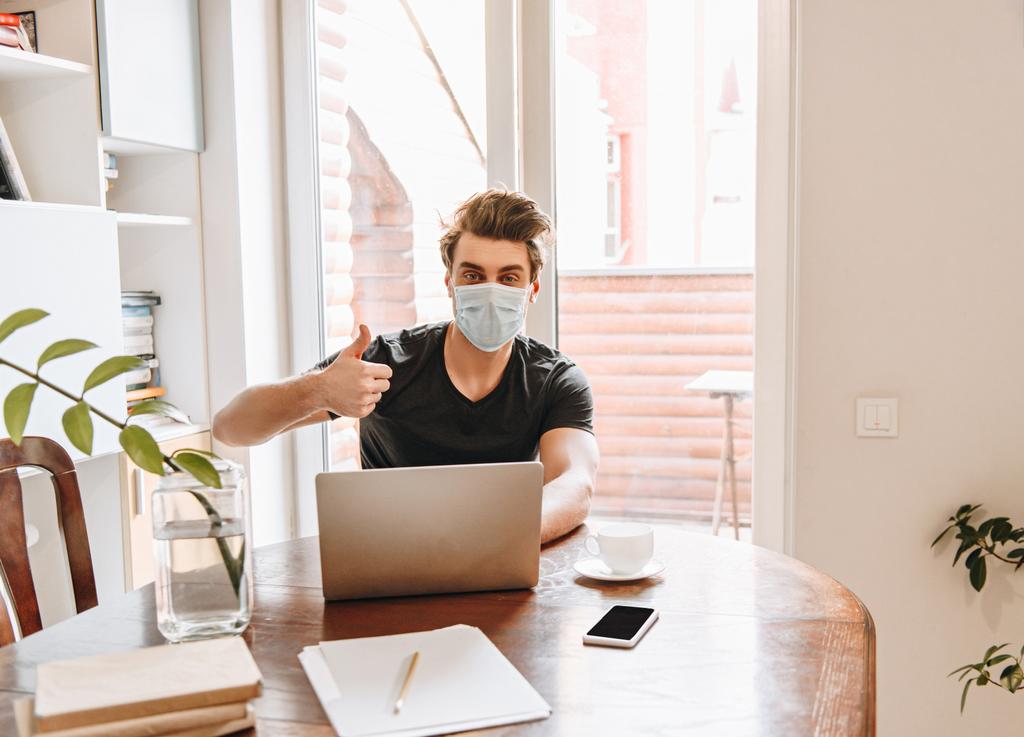 jovem homem em máscara médica mostrando polegar para cima enquanto sentado perto de laptop em casa
 - Foto, Imagem
