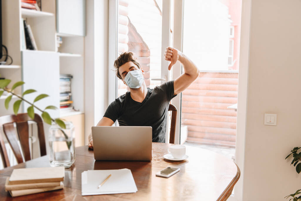 jeune homme en masque médical montrant pouce vers le bas près d'un ordinateur portable et une tasse de café
 - Photo, image