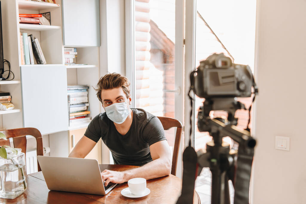 Selektiver Fokus junger Vlogger in medizinischer Maske, die in der Nähe des Laptops auf die Digitalkamera schauen - Foto, Bild