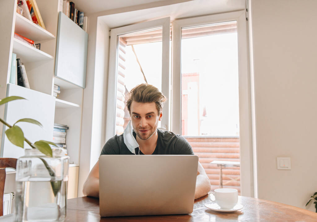 uomo sorridente con maschera medica appesa all'orecchio che lavora su laptop vicino a tazza di caffè
 - Foto, immagini