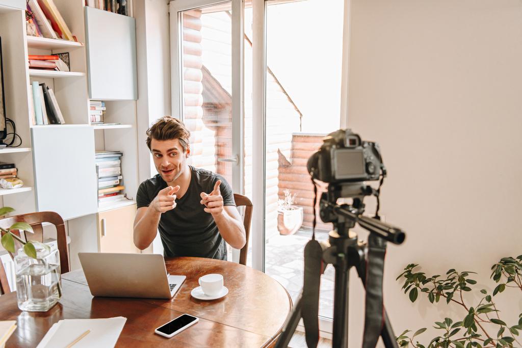 alegre vlogger apuntando con los dedos a la cámara digital mientras está sentado cerca de la computadora portátil
 - Foto, Imagen