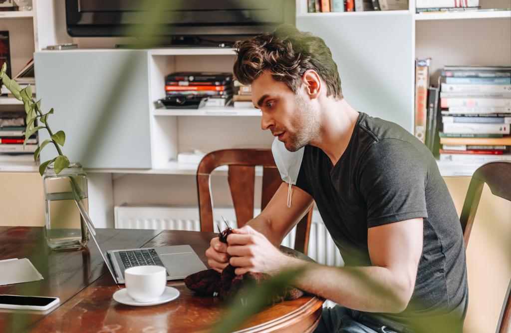 Seitenansicht eines konzentrierten jungen Mannes, der beim Stricken zu Hause auf den Laptop schaut - Foto, Bild