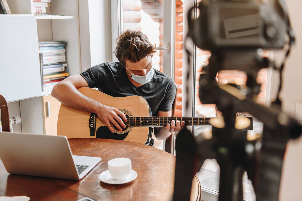 selektivní zaměření mladého vloggera v ochranné masce hrající na kytaru u notebooku a digitální fotoaparát na stativu - Fotografie, Obrázek