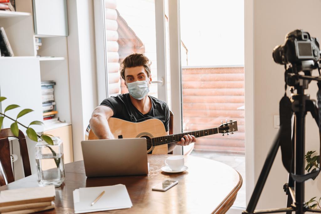 医療用マスク保持ギターの若いビデオブロガーは三脚にラップトップとデジタルカメラの近くに座っている間 - 写真・画像
