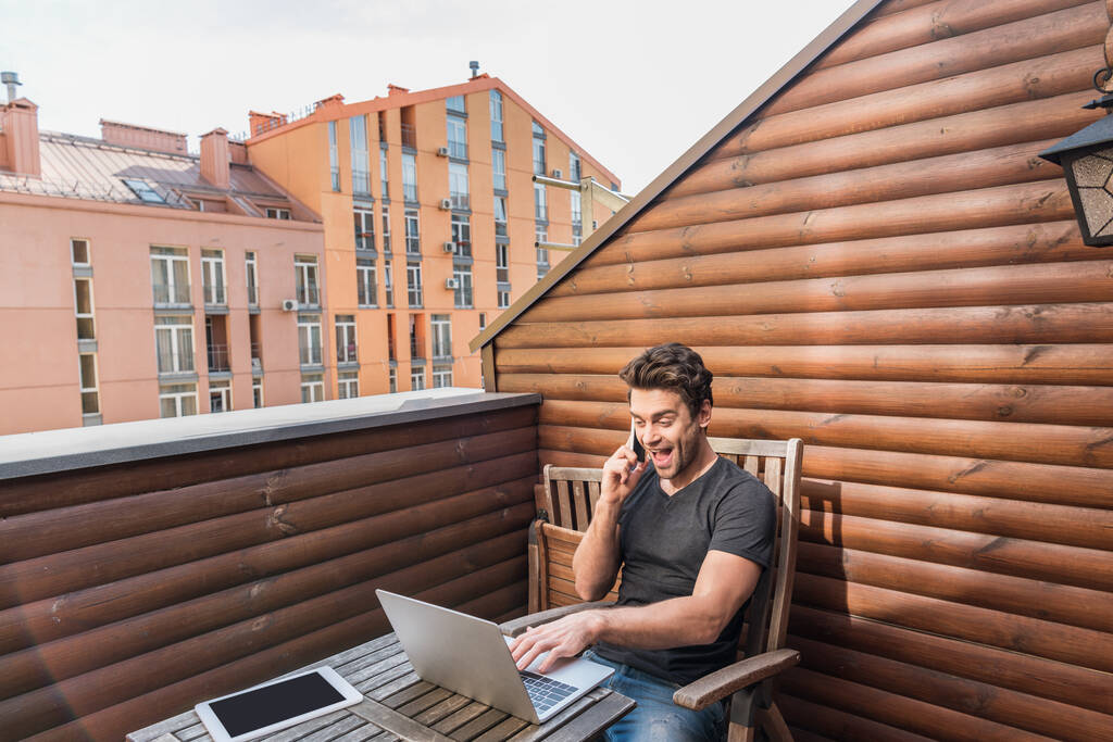 hombre emocionado usando el ordenador portátil y hablando en el teléfono inteligente mientras está sentado en el balcón
 - Foto, Imagen