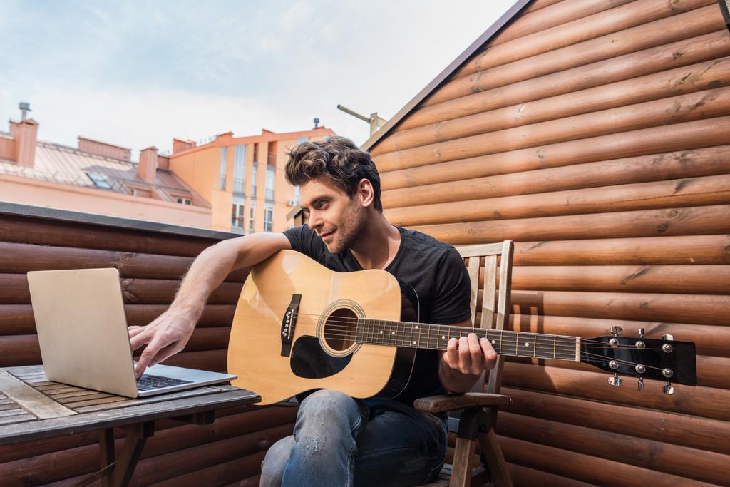 красивый молодой человек, держащий в руках гитару и ноутбук, сидя на балконе
 - Фото, изображение