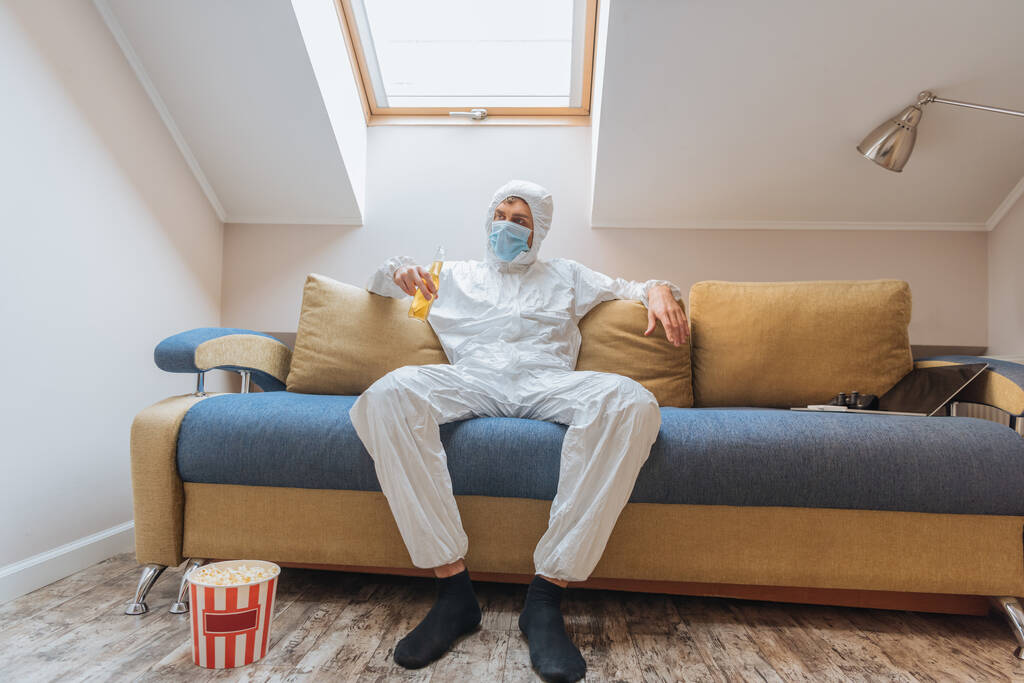 young man in hazmat suit and protective mask holding bottle of beer while sitting on sofa near popcorn bucket on floor - Photo, Image