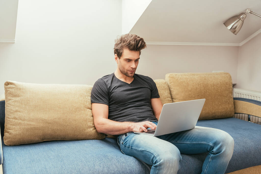 figyelmes, jóképű fiatalember laptopot használ, miközben otthon ül a kanapén. - Fotó, kép