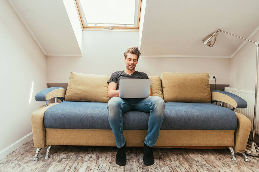 Gülümseyen genç adam evdeki kanepede otururken dizüstü bilgisayar kullanıyor. - Fotoğraf, Görsel