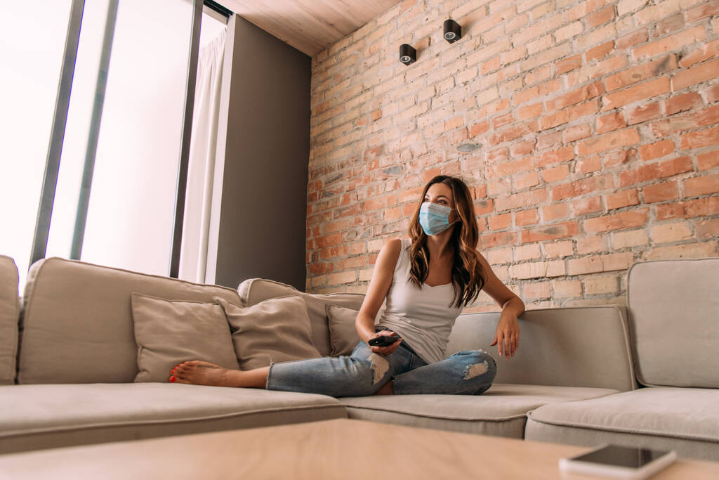 nő orvosi maszk kezében távirányító és tévénézés kanapén közben önálló elszigeteltség - Fotó, kép
