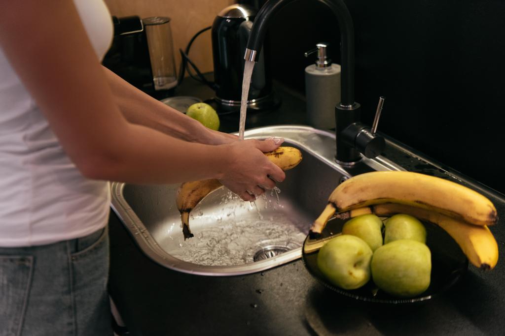 przycięty widok kobiety myjącej banany i jabłka w zlewie w kuchni podczas kwarantanny - Zdjęcie, obraz