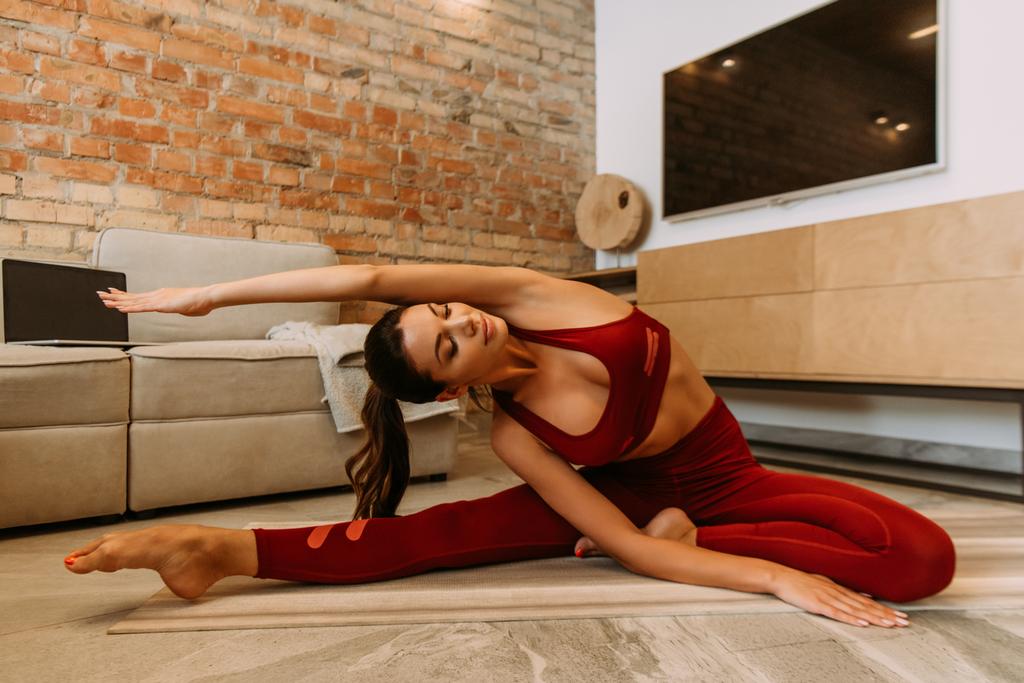 mooie vrouw stretching op yoga mat thuis met laptop op quarantaine - Foto, afbeelding