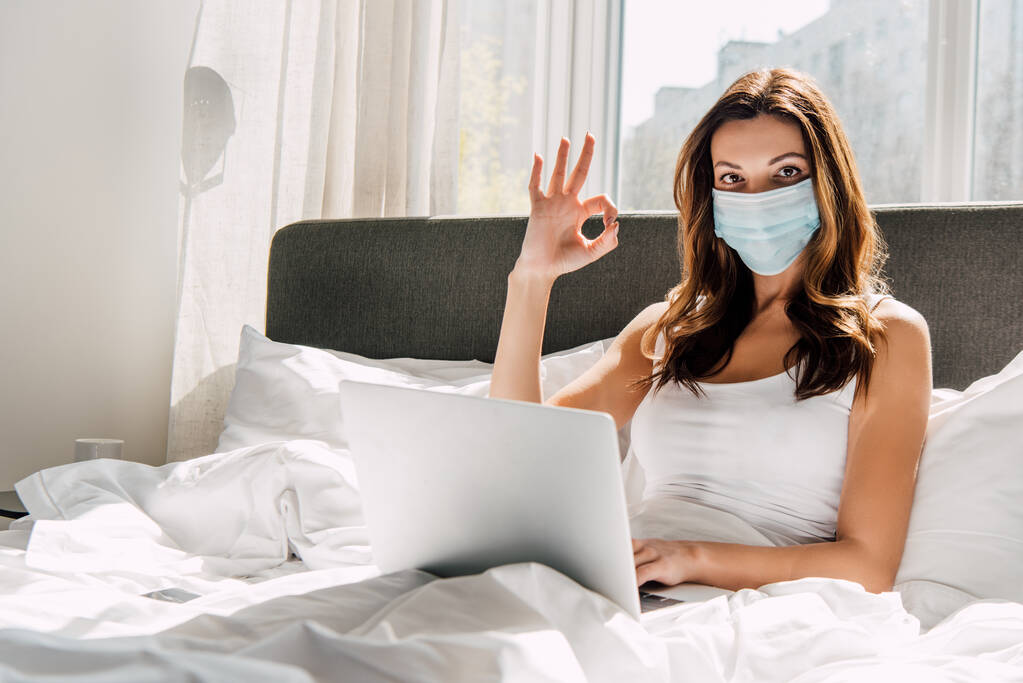 фрілансерка в медичній масці, що показує добре знак під час роботи на ноутбуці під час самоізоляції в ліжку
 - Фото, зображення