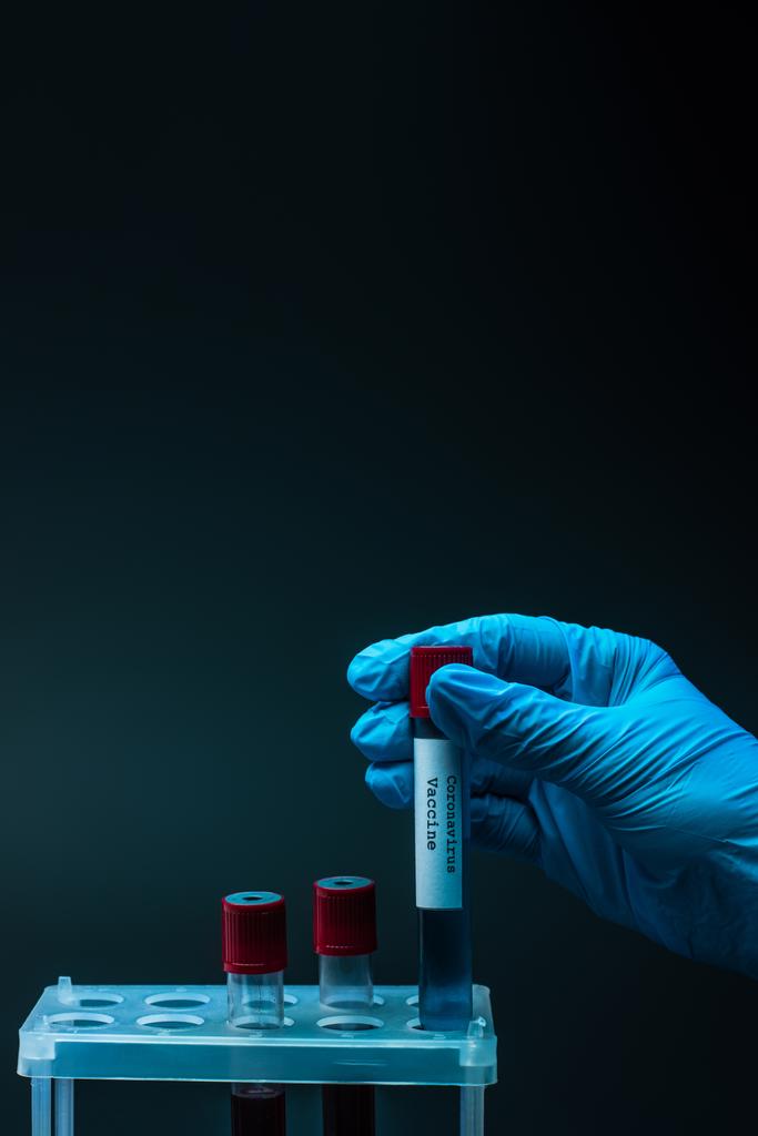 Άποψη του αρσενικού χεριού με εμβόλιο κορωναϊού και δοκιμαστικούς σωλήνες δείγματος με αίμα σε σχάρα δοκιμαστικού σωλήνα σε σκούρο φόντο - Φωτογραφία, εικόνα