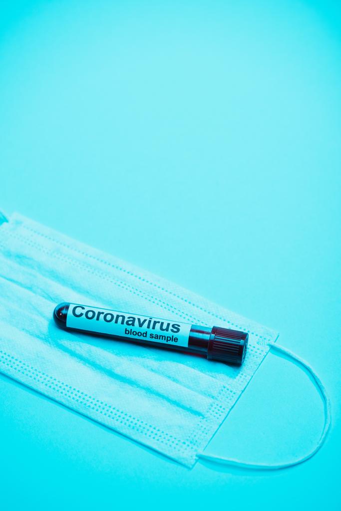 Vista de ángulo alto del tubo de ensayo con muestra de sangre de coronavirus en máscara médica sobre fondo azul
 - Foto, Imagen