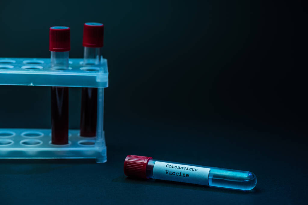 Koronavirová vakcína poblíž stojanu na zkumavky se vzorky krve na tmavé - Fotografie, Obrázek
