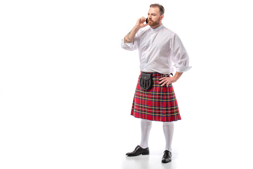 vážná skotská ryšavý muž v červeném kiltu mluvit na smartphone na bílém pozadí - Fotografie, Obrázek