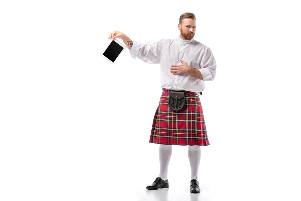 δυσαρεστημένος Σκωτσέζος κοκκινομάλλης άνδρας σε κόκκινο κιλτ με ψηφιακή ταμπλέτα σε λευκό φόντο - Φωτογραφία, εικόνα