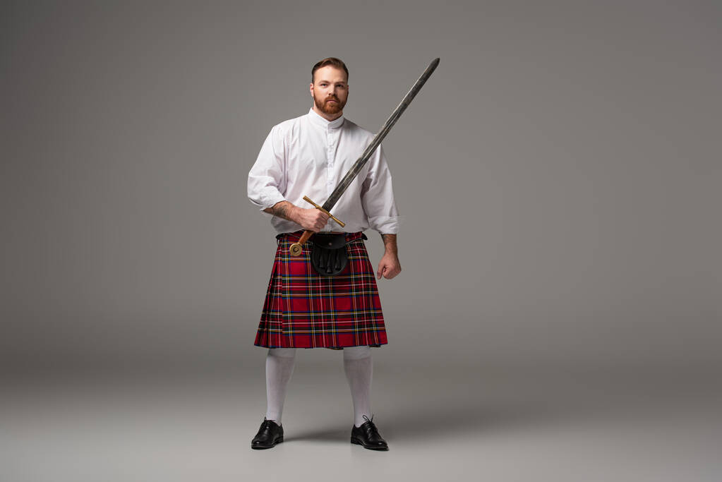 Scottish redhead man in red kilt with sword on grey background - Photo, Image