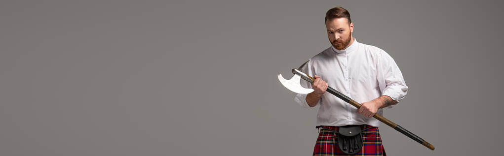 pelirrojo escocés serio en escocés rojo con hacha de batalla sobre fondo gris, tiro panorámico
 - Foto, imagen