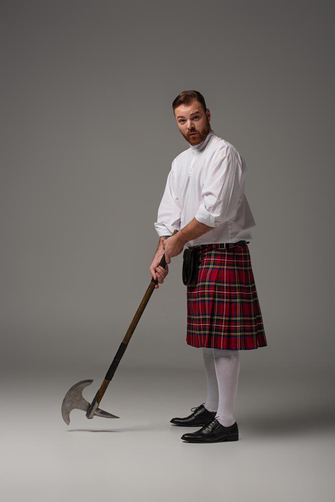 Scottish redhead man in red kilt with battle axe on grey background - Photo, Image